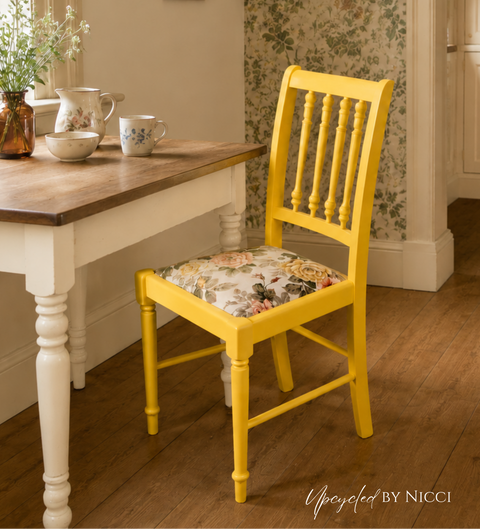 A bright yellow wooden dining chair with a floral-patterned cushion sits next to a white table with a wooden top in a cozy, vintage-style room with floral wallpaper and wood floors.