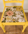 A bright yellow wooden chair with a floral upholstered seat cushion featuring large pink, yellow, and green roses on a cream background, set on a wooden floor.