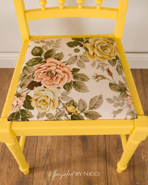 A bright yellow wooden chair with a floral upholstered seat cushion featuring large pink, yellow, and green roses on a cream background, set on a wooden floor.