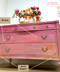 Pink dresser with gold accents in a room with flowers on a table.