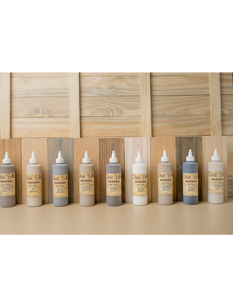 Nine bottles of Dixie Belle VooDoo Stains in varying shades are lined up on a table. The background features wooden planks in different natural tones, complementing the stains.