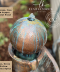 A close-up of a decorative metal garden feature, likely a fountain or sculpture, with a rounded textured top in a distressed bronze and copper finish, streaked with blue, green, and yellow patina spray. The image includes Leah Kendrick Design branding, a 'Crown Paint Company' watermark, and text labels describing the finish and colors.