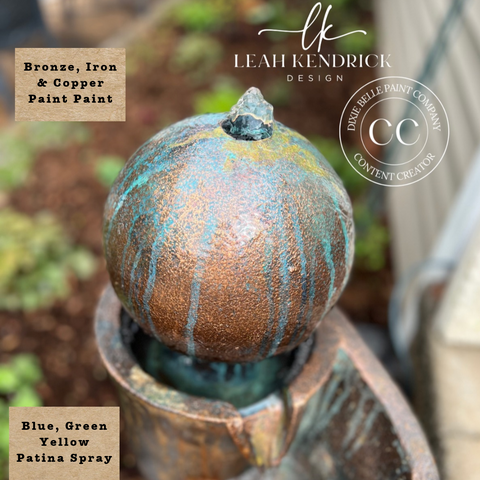 A close-up of a decorative metal garden feature, likely a fountain or sculpture, with a rounded textured top in a distressed bronze and copper finish, streaked with blue, green, and yellow patina spray. The image includes Leah Kendrick Design branding, a 'Crown Paint Company' watermark, and text labels describing the finish and colors.
