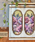 A decorative cabinet or side table with a distressed white and antique gold finish, featuring two arched floral panels with pink and purple roses. The product is shown in a styled room setting with a wooden floor, hanging greenery, and a wall, with an 'MTM Display' logo and 'Antique Gold Gilding Wax' label visible.