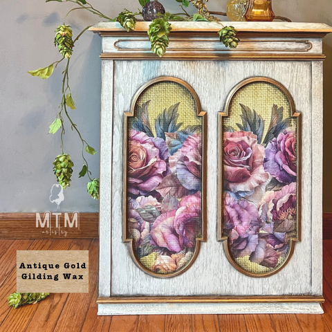 A decorative cabinet or side table with a distressed white and antique gold finish, featuring two arched floral panels with pink and purple roses. The product is shown in a styled room setting with a wooden floor, hanging greenery, and a wall, with an 'MTM Display' logo and 'Antique Gold Gilding Wax' label visible.