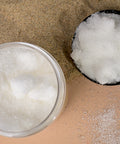 Top-down view of a jar and a black lid, both filled with whitish clear granules, placed on a beige surface sprinkled with sand and white granules.