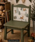 A rustic children's wooden chair with a distressed sage green finish and a winter woodland decoupage paper on the backrest showing bears, squirrels, and snow trees. The chair is styled in a cozy room setting with a teddy bear, books, a basket, and a potted evergreen tree.