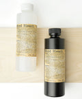 Two bottles labeled "Wood Bleach" are placed on a light wooden board. One bottle is clear with a white cap, and the other is black with a black cap. Both have beige labels with detailed text.