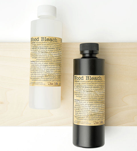 Two bottles labeled "Wood Bleach" are placed on a light wooden board. One bottle is clear with a white cap, and the other is black with a black cap. Both have beige labels with detailed text.