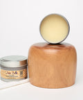 An open jar of Dixie Belle wood conditioner is placed in front of a wooden block and a polished wooden object. A white cloth rests beside the jar, all set against a plain white background.