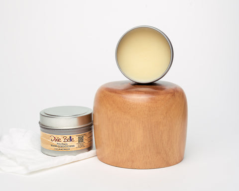 An open jar of Dixie Belle wood conditioner is placed in front of a wooden block and a polished wooden object. A white cloth rests beside the jar, all set against a plain white background.