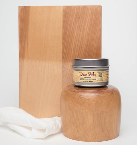 A jar of Dixie Belle wood conditioner is placed in front of a wooden block and a polished wooden object. A white cloth rests beside the jar, all set against a plain white background.