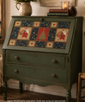 A rustic green secretary desk cabinet decorated with a Christmas-themed patchwork panel on the front. The cabinet features two drawers with round brass knobs and stands on turned legs.