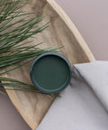 Overhead image of an open jar of Acadia. Staged on a wooden bowl with faux greenery and a drop cloth.