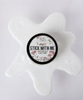 A container of “Stick With Me” Dixie Shine glue sits on a white surface, surrounded by a splatter-shaped spill of glue. The round label features decorative floral accents.