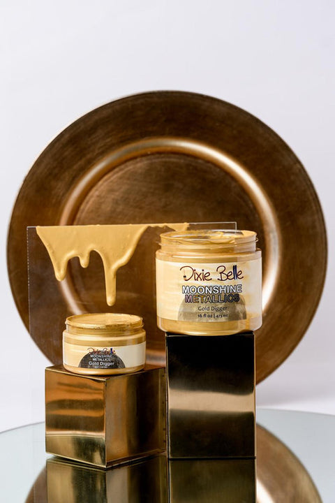 Two jars of Dixie Belle Moonshine Metallics "Gold Digger" paint are displayed on gold cubes, with gold paint dripping on clear acrylic behind them, and a large gold plate in the background.