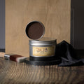A container of Dixie Belle Best Dang Wax, open to show brown wax inside. It is placed on a wooden surface with a wooden backdrop, accompanied by a paintbrush and black cloth.