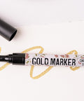 A gold marker with a chisel tip lies on a white surface. The cap is removed and placed nearby. The marker has a floral design on its body, and a gold line is drawn on the surface.