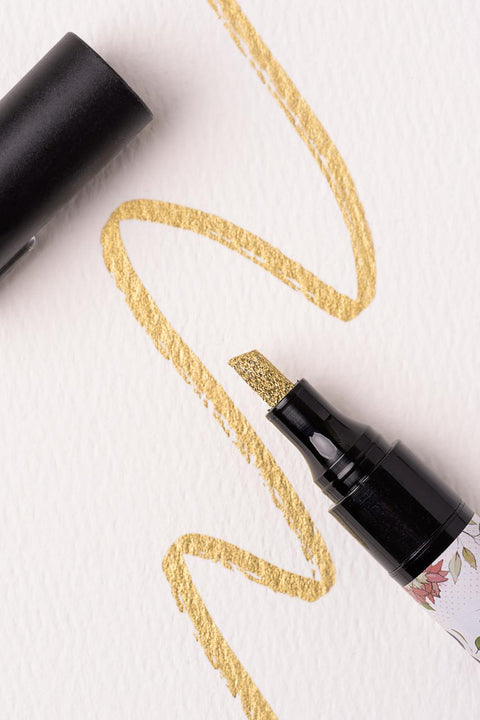A black marker with a gold glitter tip is shown with the cap off. The marker is drawing a wavy gold line on textured white paper. The design on the marker barrel includes floral patterns.