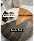 A round wooden board shows three sections with different wood stains: Picklin White, American Honey, and Colonial Black. A cup of black grapes and a small wooden apple rest on the board. Containers labeled Flour and Sugar are in the background.