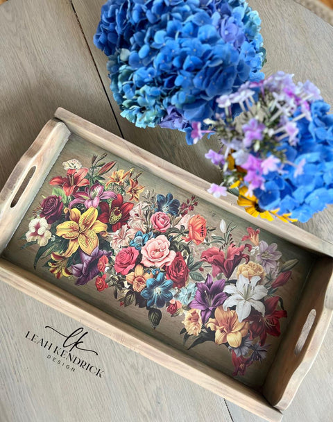 A wooden tray on a table features a vibrant array of flowers, including roses, lilies, and other blooms. Next to the tray, a bouquet of blue and purple hydrangeas adds a touch of freshness. "Leah Kendrick Design" watermark logo placed below the tray.
