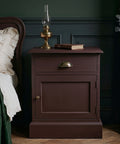 Dark moody bedroom featuring a vintage-style wooden nightstand painted in a rich red-brown finish, styled with a brass oil lamp and book beside a bed with cream bedding and deep green accents.