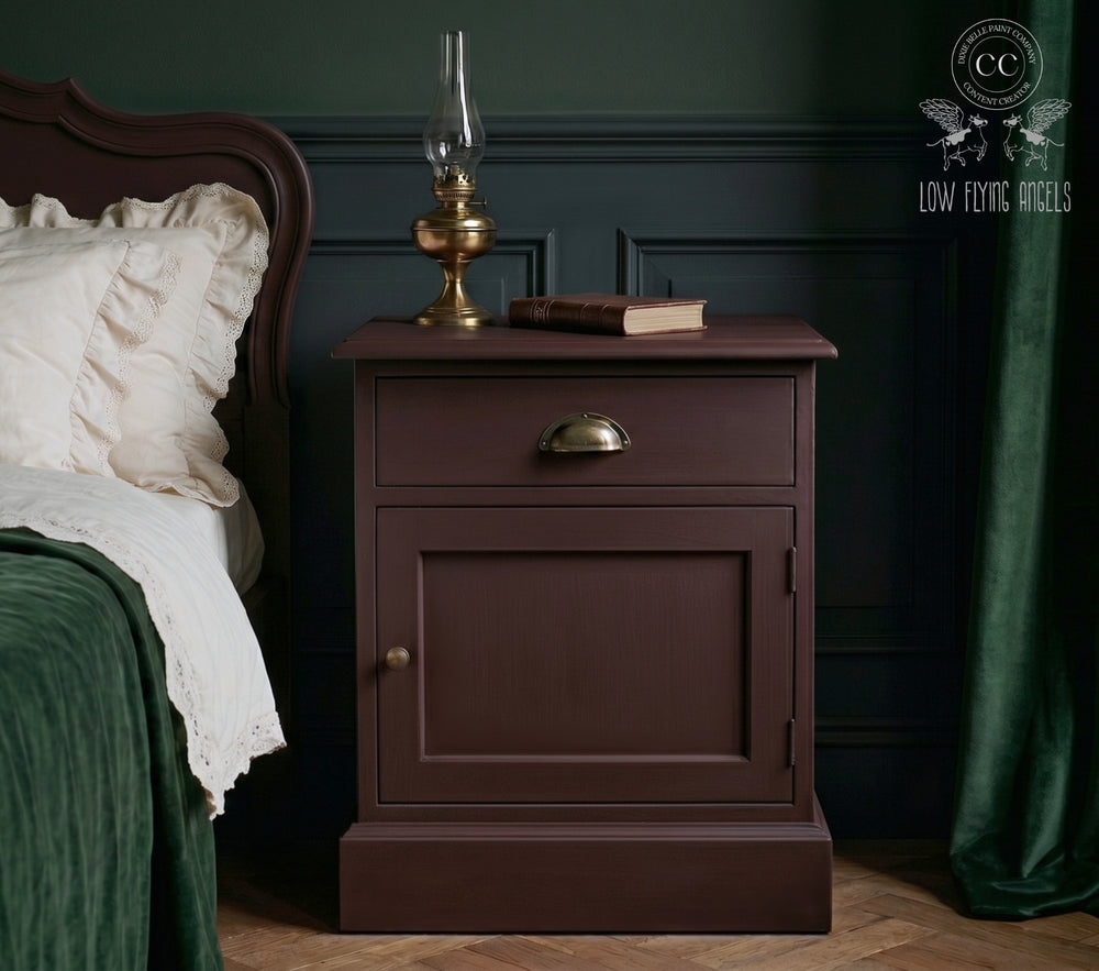 Dark moody bedroom featuring a vintage-style wooden nightstand painted in a rich red-brown finish, styled with a brass oil lamp and book beside a bed with cream bedding and deep green accents.