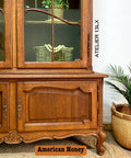 A vintage wooden hutch with ornate details sits beside a basket holding a potted plant. A woven basket with greenery is displayed inside the hutch. Text reads “ATELIER 13LX” and “American Honey.”.