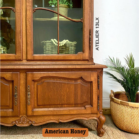A vintage wooden hutch with ornate details sits beside a basket holding a potted plant. A woven basket with greenery is displayed inside the hutch. Text reads “ATELIER 13LX” and “American Honey.”.