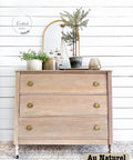 A rustic wooden dresser with three drawers features gold knobs, sitting against a white shiplap wall. It is topped with a round mirror, a candelabra, and various potted plants. The text "Au Naturel" is in the bottom-right corner.