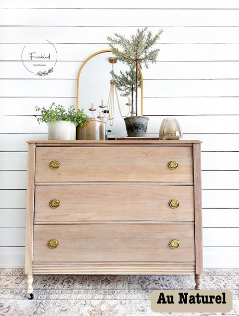 A rustic wooden dresser with three drawers features gold knobs, sitting against a white shiplap wall. It is topped with a round mirror, a candelabra, and various potted plants. The text "Au Naturel" is in the bottom-right corner.