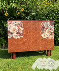 A wooden chest sits on a grassy lawn in front of tall green plants and yellow flowers. The chest is decorated with intricate patterns in red and gold, and features floral designs of large blossoms on its top corners. Two small certification badges are visible at the bottom.