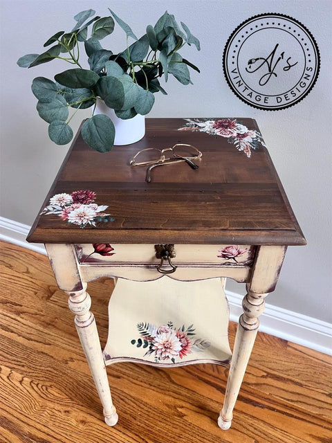 A small square wooden side table with a dark brown top featuring floral designs in white, pink, and red. The table has a single drawer with matching floral artwork and a lower shelf. A potted plant and glasses rest on the table. "AJs Vintage Designs" logo is visible.