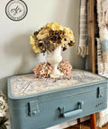 Belles and Whistles Ancient Marrakesh - A1 Rice Decoupage Paper. Beautiful light blue suitcase end table with Ancient Marrakesh decoupage on top. The piece is staged with a yellow and pink flower arrangement against a white wall.