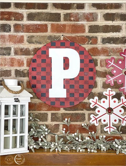 Belles and Whistles Buffalo Check - Stencil provides an extra decorative touch to any project!
A red round sign with a large white letter "P" on the background has Buffalo Check - Stencil in black. Presented on a red brick wall hanging above a row of mistletoe and a  white and red snowflake signs is seen on the right.