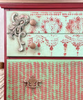 Belles and Whistles Cozy Sweater - Stencil provides an extra decorative touch to any project! A closeup image of a dresser that is painted in 2 colors with a mauve pink being on the sides and then a light blue on the drawer fronts. The first drawer has our lotus bloom stencil in the same mauve color and the second drawer features our cozy sweater stencil.