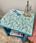 Belles and Whistles Enchanted Garden - Stencil provides an extra decorative touch to any project! Blue painted end table with the Enchanted Garden Stencil design applied on top with a dragonfly. Staged with a clear mason jar on a wooden coaster on top.