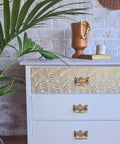Belles and Whistles Enchanted Garden - Stencil provides an extra decorative touch to any project! White painted dresser with gold handles. Enchanted Garden Stencil applied on the top drawer front in gold. Staged with a book with gold pages on top and an orange vase with wooden beads spilling out of it.