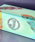 Belles and Whistles Feathers - Rice Decoupage Paper 
Here is a a plain wooden box painted in a light green and has an iron, patinaed "J" with a metal circle around the "J" on the front of the box.  The box can be opened like a jewelry box.  It has some of the Belles and Whistles decoupage feathers used on the box.  It is staged on a purple surface with the same purple behind the box.  There is a yellow planter with a green plant and white pebbles in front of the left side of the box.