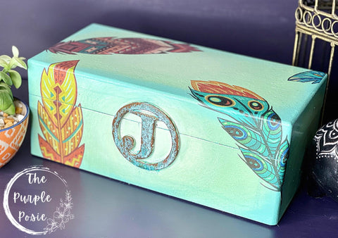 Belles and Whistles Feathers - Rice Decoupage Paper 
Here is a a plain wooden box painted in a light green and has an iron, patinaed "J" with a metal circle around the "J" on the front of the box.  The box can be opened like a jewelry box.  It has some of the Belles and Whistles decoupage feathers used on the box.  It is staged on a purple surface with the same purple behind the box.  There is a yellow planter with a green plant and white pebbles in front of the left side of the box.