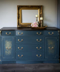 Belles and Whistles Greek Key - Stencil provides an extra decorative touch to any project! Dresser painted in a muted navy blue  with gold hardwares and Greek Key - Stencil  in gold faded a bit on the doors of the piece. Staged with a gold frame on the center of the dresser , with gold tray and two candle holder on top with some flowers.