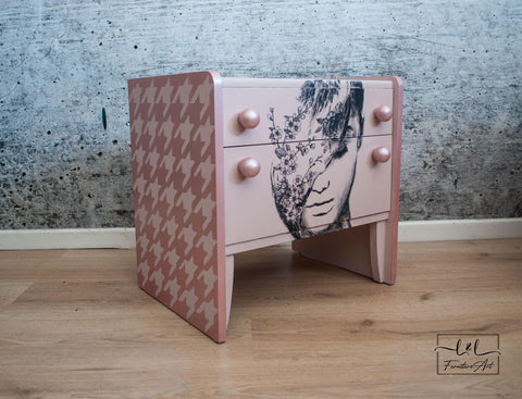 Belles and Whistles Houndstooth - Stencil provides an extra decorative touch to any project!
A light pink bedside dresser with the side panels covered in Houndstooth - Stencil with a lighter pink. Staged with laminated wood floor and a gray concrete looking wall.