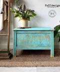 Belles and Whistles Lotus Bloom - Stencil provides an extra decorative touch to any project!
Coffee table painted a teal blue with Lotus Bloom - Stencil in gold along the middle panel with gold accent on the stencil. Staged on a woven carpet , a flower pot with greenery and flowers.