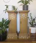Belles and Whistles Lotus Bloom - Stencil provides an extra decorative touch to any project!
Two tan slender cabinet pillars with Lotus Bloom - Stencil in white at the top and base of furniture. Staged with flower pots on each side and on top of both pieces with greenery.