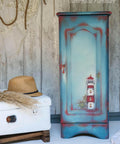 Belles and Whistles Nautical - Silkscreen Stencil provides an extra decorative touch to any project! Blue and red chalk painted armoire with a light house and ship wheel stenciled on. Staged next to a white luggage.