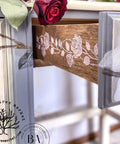 Belles and Whistles Roses - Silkscreen Stencil provides an extra decorative touch to any project! A closeup of the side of the drawers on a desk. The vertical rose design in white is shown on the the drawer side, staged with roses on top of the desk.