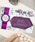 Belles and Whistles Thing-a-ma-jig  - Thing-a-ma-jig in it's packaging sitting on a gray and ivory striped table with ivory beads and green & purple leaves around.