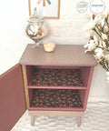 Belles and Whistles Trippy Blocks - Stencil.  A pink chalk painted night stand with the front door open.  The inside shelves are covered with pink and black floral paper.  the top of the nightstand has the Trippy Blocks stencil applied in tan chalk paint.  The base and legs of the table are painted with tan chalk paint.  Staged on white carpet, in front of a white wall.  White flowers are to the right of the night stand, and a glass vase holding flowers and a white candle sit on top of the night stand.