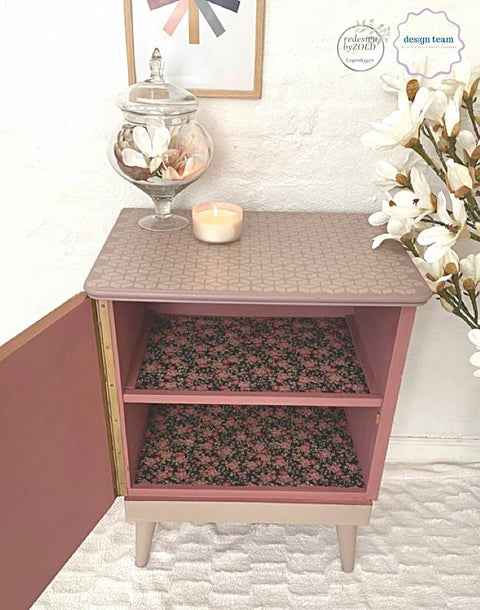 Belles and Whistles Trippy Blocks - Stencil.  A pink chalk painted night stand with the front door open.  The inside shelves are covered with pink and black floral paper.  the top of the nightstand has the Trippy Blocks stencil applied in tan chalk paint.  The base and legs of the table are painted with tan chalk paint.  Staged on white carpet, in front of a white wall.  White flowers are to the right of the night stand, and a glass vase holding flowers and a white candle sit on top of the night stand.