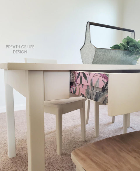 Belles and Whistles Tropical on Pink - Rice Decoupage Paper.  A white chalk painted table with the drawer partially open.  the side of the drawer has the Tropical on Pink Rice Decoupage Paper applied.  Staged on off-white carpet with a white chair and a silver metal tray holing greenery on top of the table.